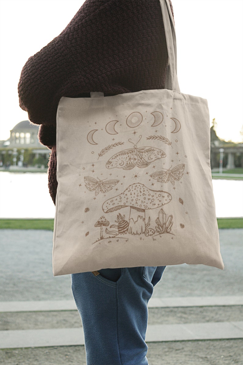 May include: A beige tote bag with a brown design of a mushroom, butterflies, and moons. The design is reminiscent of a mystical forest scene.