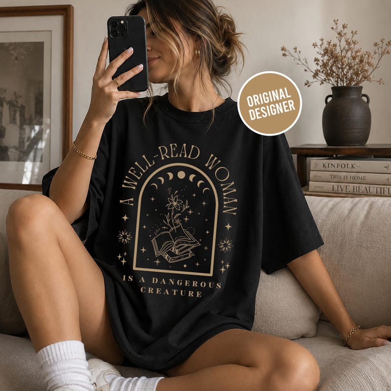 K&ouml;nnte beinhalten: Schwarzes T-Shirt mit goldenem Aufdruck: &bdquo;A WELL-READ WOMAN IS A DANGEROUS CREATURE&ldquo;. Das Design zeigt einen Bogen mit Monden, Sternen und einem offenen Buch. Das Shirt hat eine lockere, &uuml;bergro&szlig;e Passform.