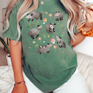 May include: A green t-shirt with a graphic print of multiple opossums in a forest setting. The opossums are depicted in various poses, including holding a baby opossum, sitting on a mushroom, and walking through the leaves. The graphic is surrounded by flowers, leaves, and mushrooms.
