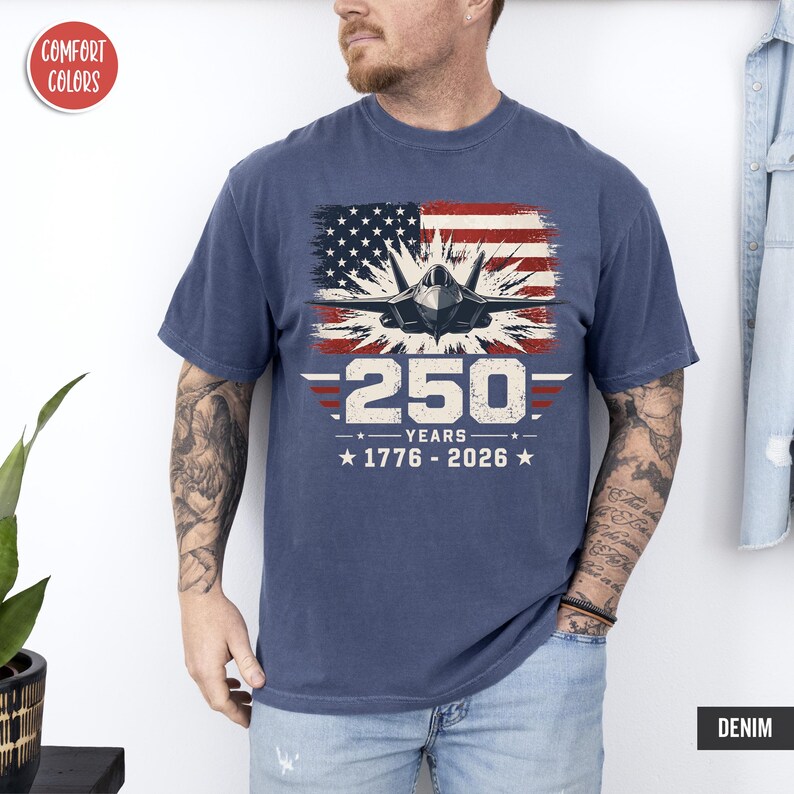 May include: Denim blue t-shirt featuring a graphic of a fighter jet in front of an American flag. The text reads "250 YEARS 1776-2026". The shirt is a Comfort Colors brand.