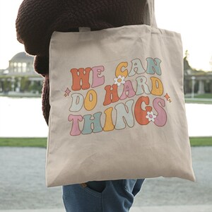 Op de afbeelding: Een beige canvas draagtas met de tekst "WE CAN DO HARD THINGS" in kleurrijke retro-stijl letters met bloemen.