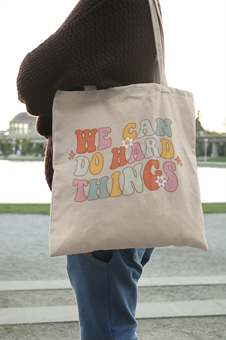 Op de afbeelding: Een beige canvas draagtas met de tekst "WE CAN DO HARD THINGS" in kleurrijke retro-stijl letters met bloemen.
