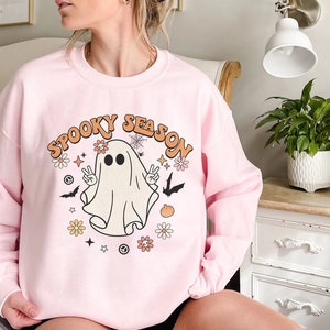 May include: Pink sweatshirt with a white ghost graphic and the text "Spooky Season" in a retro font. The ghost has two eyes and is surrounded by flowers, bats, and stars.