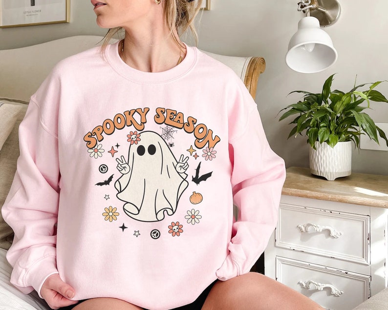 May include: Pink sweatshirt with a white ghost graphic and the text "Spooky Season" in a retro font. The ghost has two eyes and is surrounded by flowers, bats, and stars.