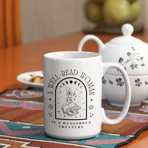 Pode incluir: Caneca de cer&acirc;mica branca com um design preto que diz "Uma mulher bem lida &eacute; uma criatura perigosa". O design apresenta um livro com uma flor crescendo dele, rodeado de estrelas e luas.