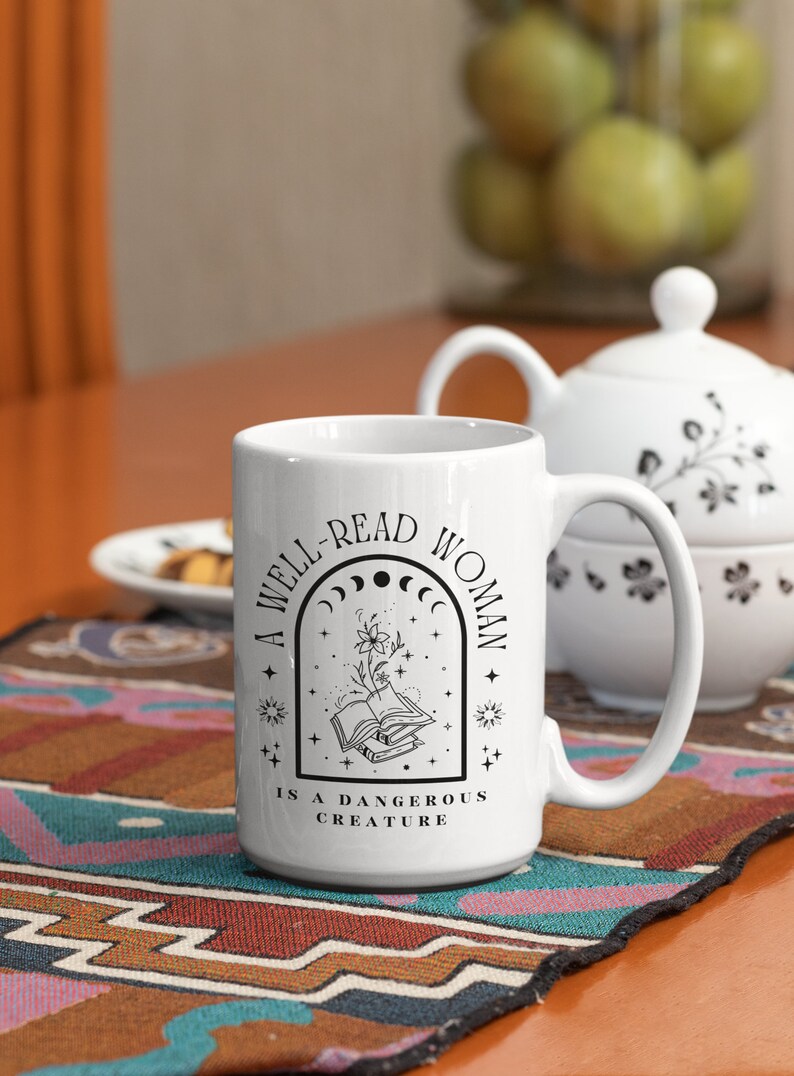 Pode incluir: Caneca de cer&acirc;mica branca com um design preto que diz "Uma mulher bem lida &eacute; uma criatura perigosa". O design apresenta um livro com uma flor crescendo dele, rodeado de estrelas e luas.
