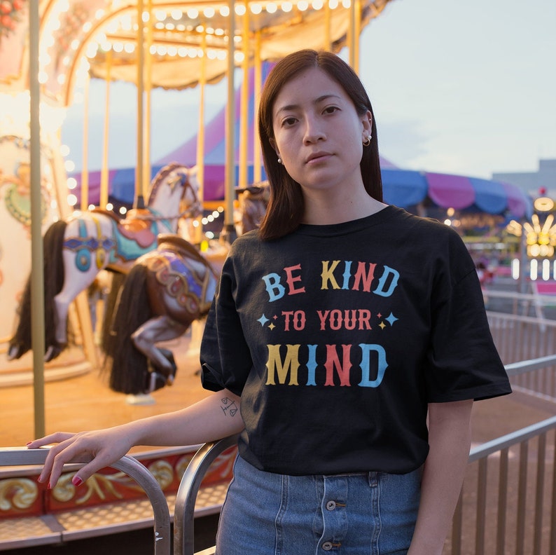 May include: A person wearing a black t-shirt with the text "BE KIND TO YOUR MIND" in colorful letters. The person is standing in front of a carousel.