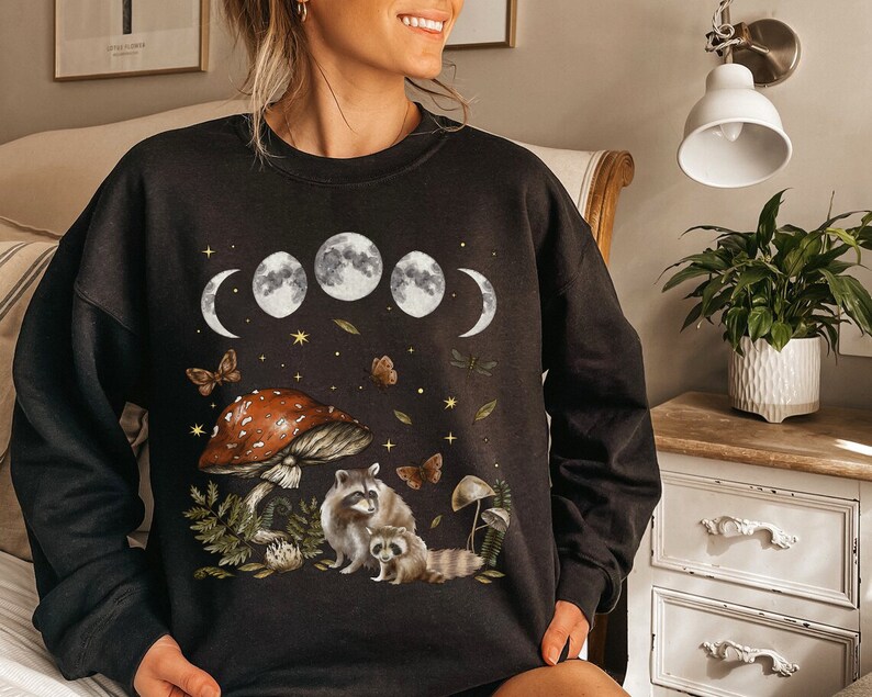 May include: Black sweatshirt with a graphic print of raccoons, mushrooms, and a crescent moon phase. The graphic is in shades of brown, white, and gray.