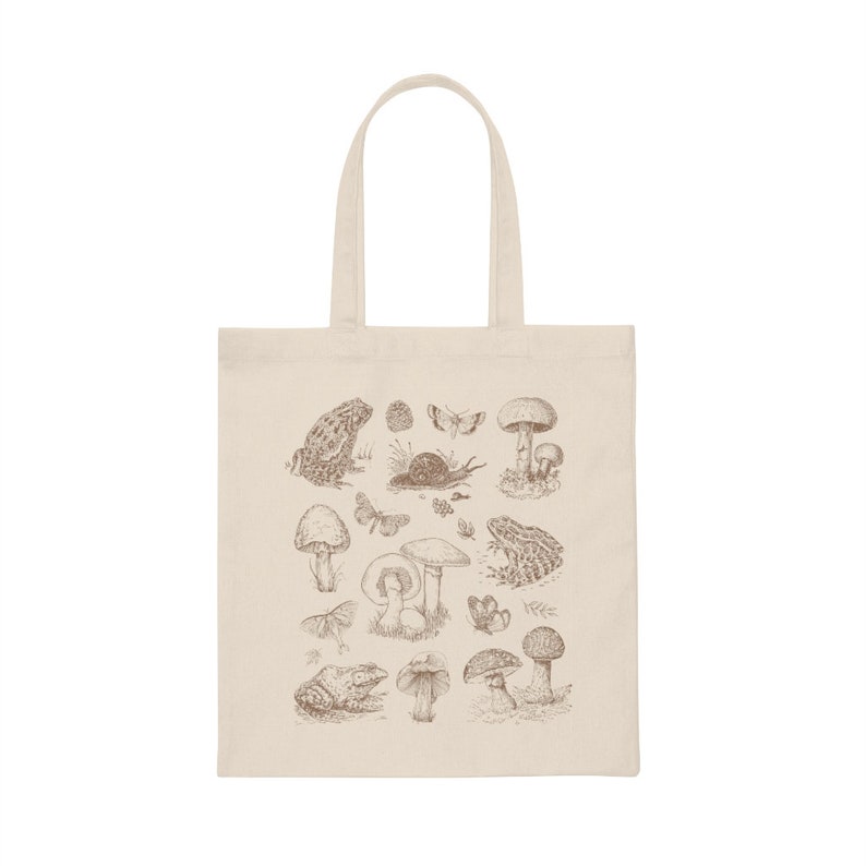May include: A beige tote bag with a brown illustration of mushrooms, frogs, butterflies, and snails. The design is centered on the bag.