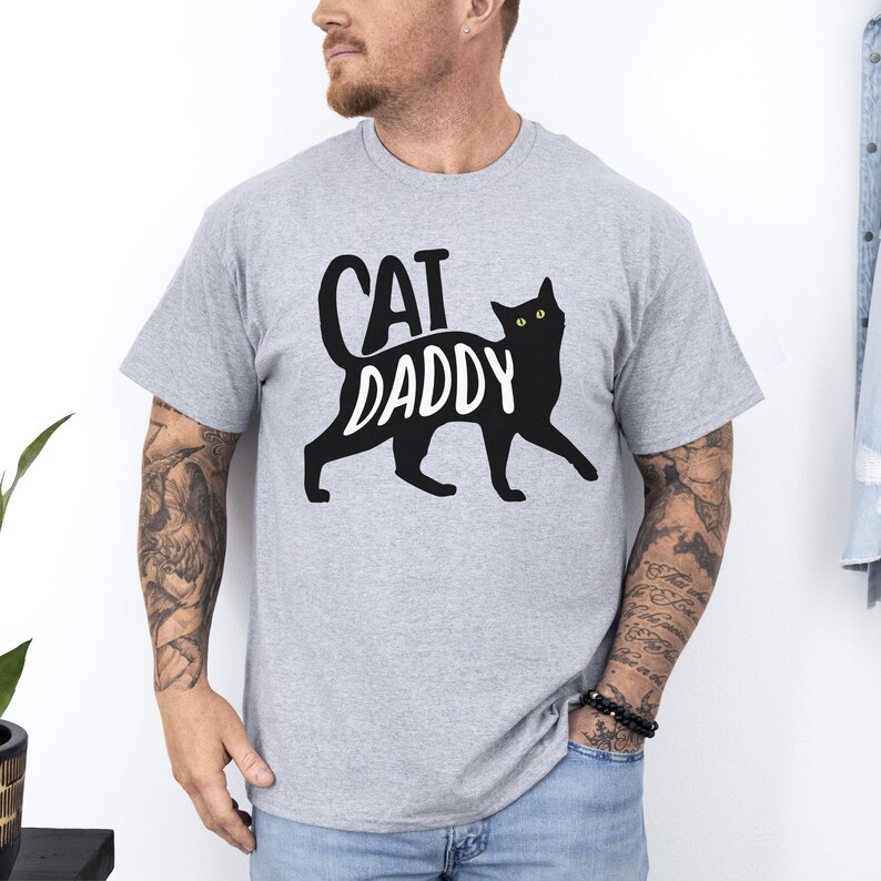 May include: A light gray t-shirt with the words "CAT DADDY" in black, featuring a black cat silhouette. The cat has yellow eyes. The shirt is short-sleeved.