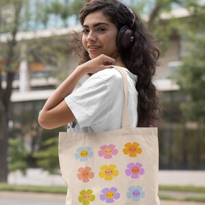 Pu&ograve; includere: Una borsa tote beige con un motivo floreale colorato e il testo "think happy thoughts". I fiori hanno facce sorridenti e sono in tonalit&agrave; di rosa, giallo, blu, verde e arancione.