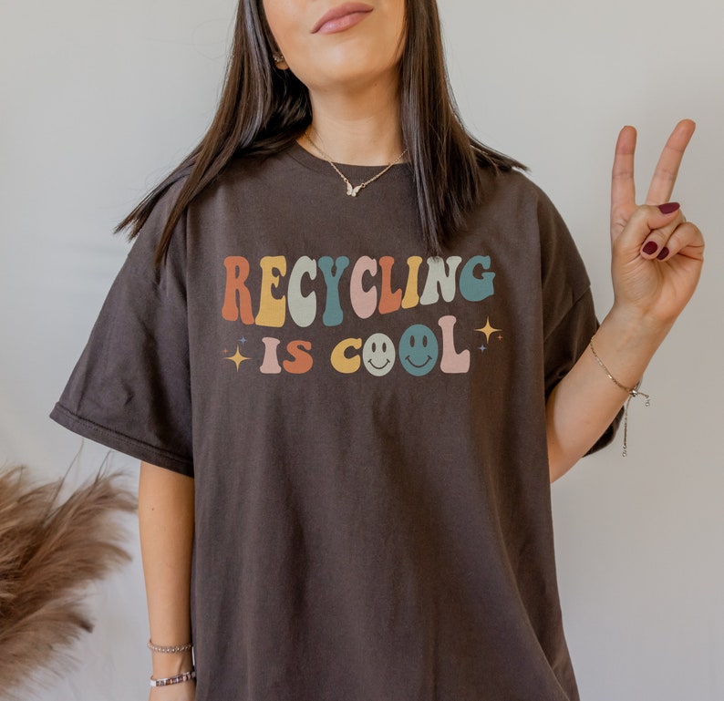K&ouml;nnte beinhalten: Ein braunes T-Shirt mit dem Text "Recycling is cool" in bunten Retro-Buchstaben mit einem Smiley.
