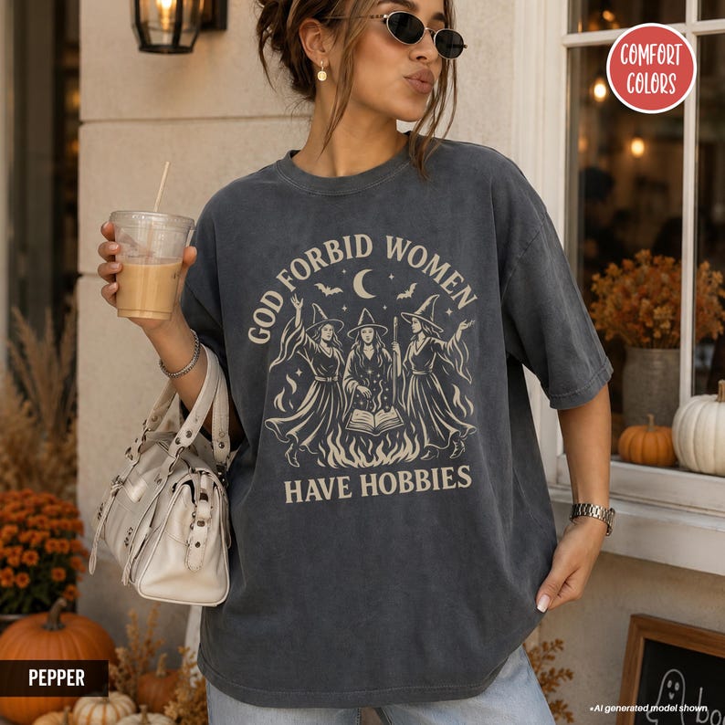 May include: A charcoal gray short-sleeve t-shirt featuring a graphic print of three witches standing around an open book and a fire. The text reads GOD FORBID WOMEN HAVE HOBBIES. The design includes stars, a crescent moon, and bats.