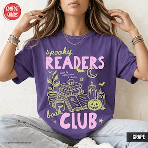 May include: A grape purple t-shirt featuring the text "spooky READERS book CLUB" in pink and "fueled by coffee, horror & chapters" in white. The graphic includes a stack of books, a skull, a pumpkin, candles, a plant, and a crescent moon.