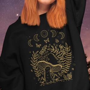 Op de afbeelding: Zwart sweatshirt met een gouden omtrek van een paddenstoel, maanfasen, vlinders en gebladerte. Het ontwerp heeft een speelse, door de natuur ge&iuml;nspireerde esthetiek.