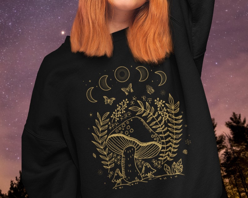 Op de afbeelding: Zwart sweatshirt met een gouden omtrek van een paddenstoel, maanfasen, vlinders en gebladerte. Het ontwerp heeft een speelse, door de natuur ge&iuml;nspireerde esthetiek.