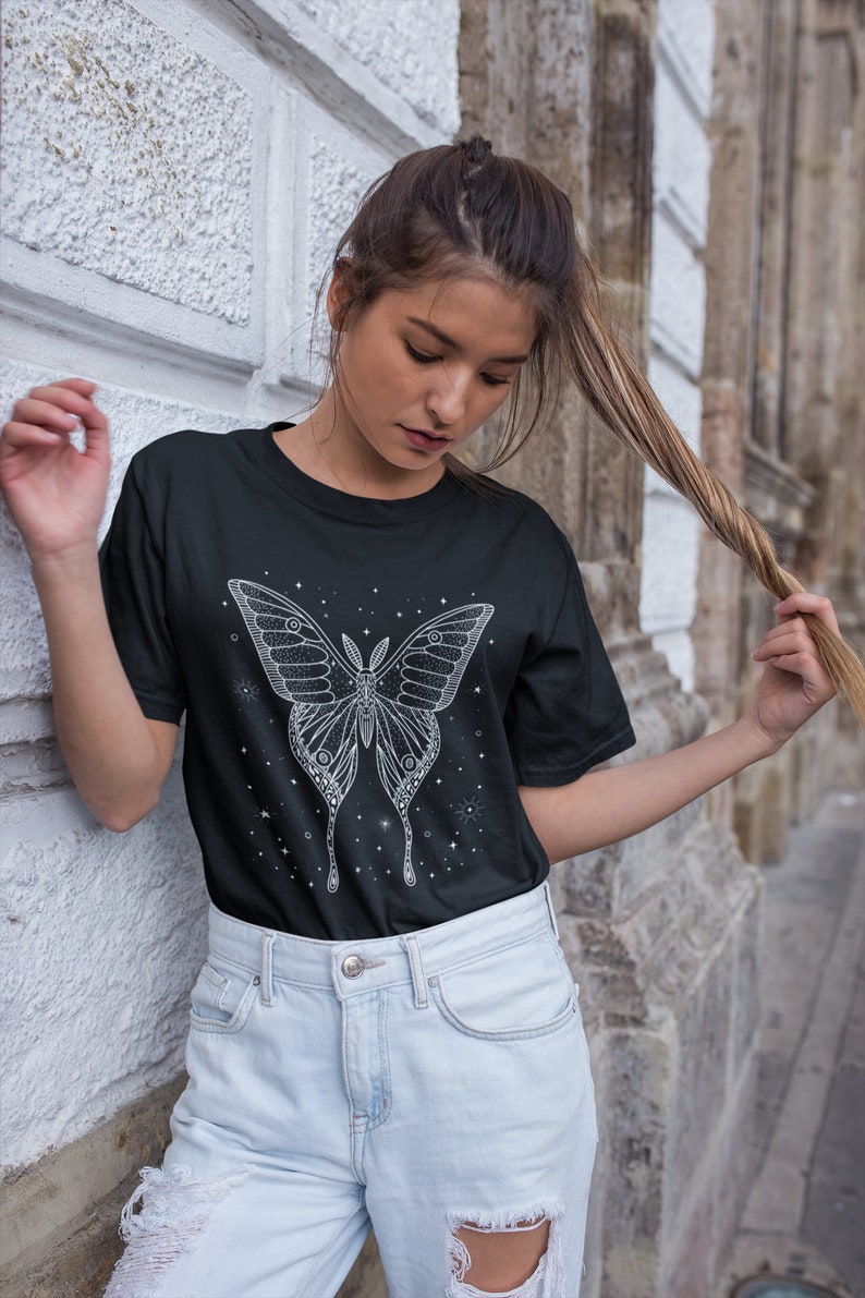 May include: A black t-shirt with a white graphic of a butterfly with stars. The butterfly has intricate details and is surrounded by small stars.