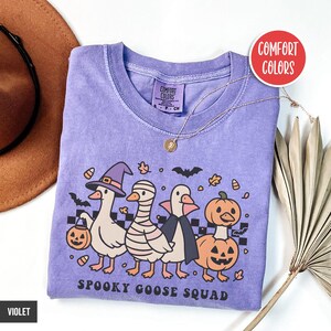 May include: A violet Comfort Colors t-shirt with a Halloween design of four cartoon geese in costume: witch, mummy, vampire, and pumpkin. The text "Spooky Goose Squad" is below. A gold necklace is draped across the shirt. The shirt is violet.