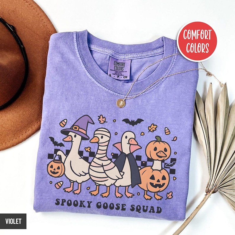 May include: A violet Comfort Colors t-shirt with a Halloween design of four cartoon geese in costume: witch, mummy, vampire, and pumpkin. The text "Spooky Goose Squad" is below. A gold necklace is draped across the shirt. The shirt is violet.