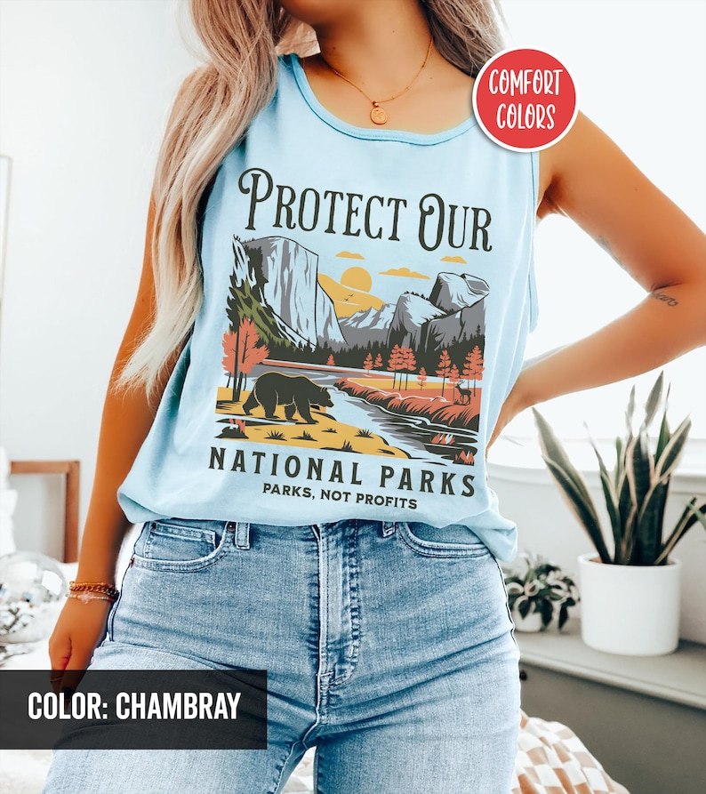 May include: Light blue tank top with a graphic that reads "PROTECT OUR NATIONAL PARKS". The graphic features a bear, mountains, and a river. The text "PARKS, NOT PROFITS" is also included. The tank top is labeled "COMFORT COLORS" and "COLOR: CHAMBRAY".