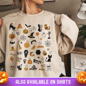 May include: A beige sweatshirt with a Halloween-themed design featuring a variety of spooky and cute icons, including pumpkins, ghosts, bats, cats, and candy corn. The text "BOO!" is also included in the design.