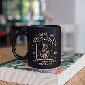 May include: Black ceramic mug with the text "A WELL-READ WOMAN IS A DANGEROUS CREATURE" in a cream-colored font. The mug features a design of an open book with floral elements and celestial motifs. The mug is resting on a book with a colorful cover.