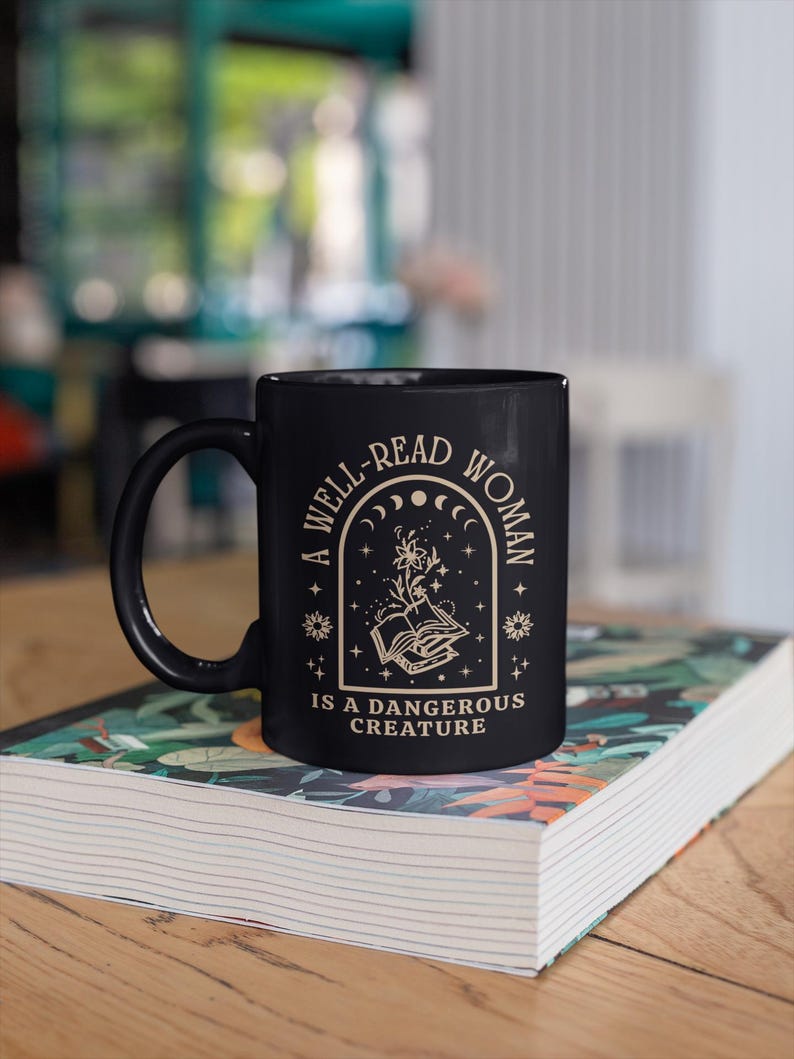 May include: Black ceramic mug with the text "A WELL-READ WOMAN IS A DANGEROUS CREATURE" in a cream-colored font. The mug features a design of an open book with floral elements and celestial motifs. The mug is resting on a book with a colorful cover.