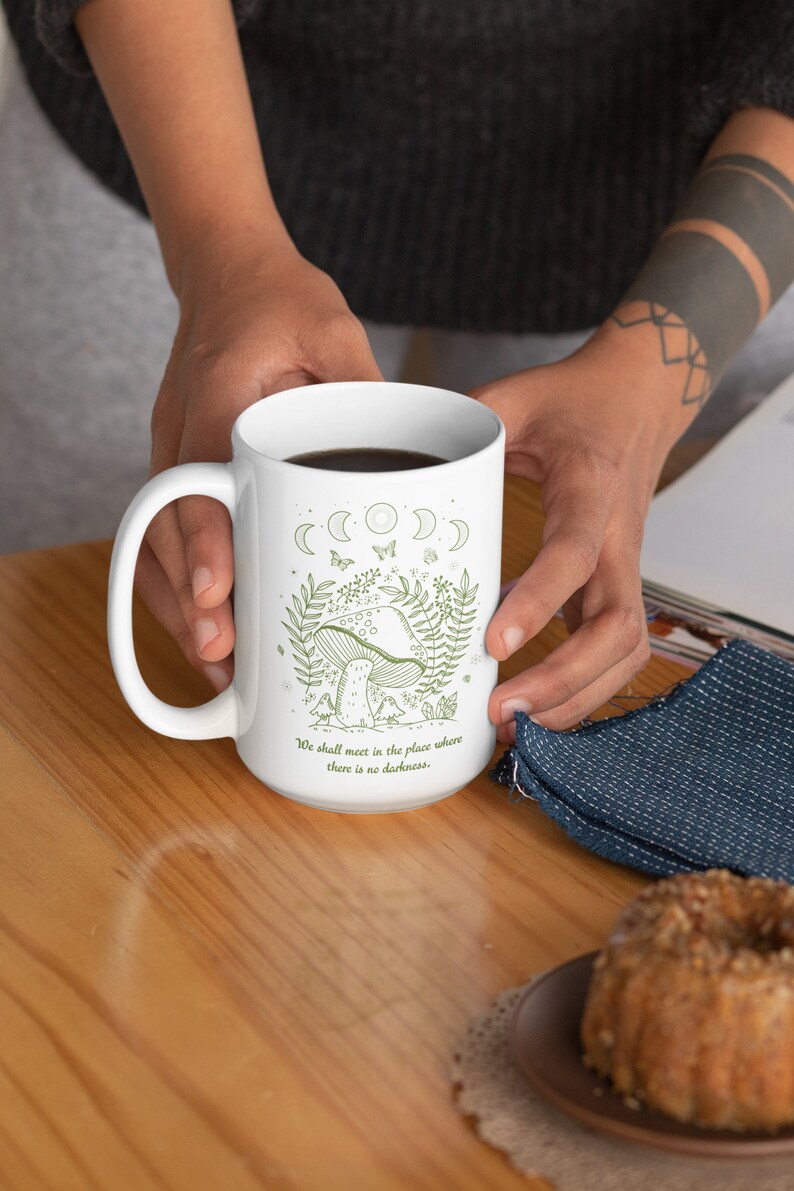 May include: White ceramic mug with a green illustration of a mushroom, moon phases, butterflies, and the text "We shall meet in the place where there is no darkness."