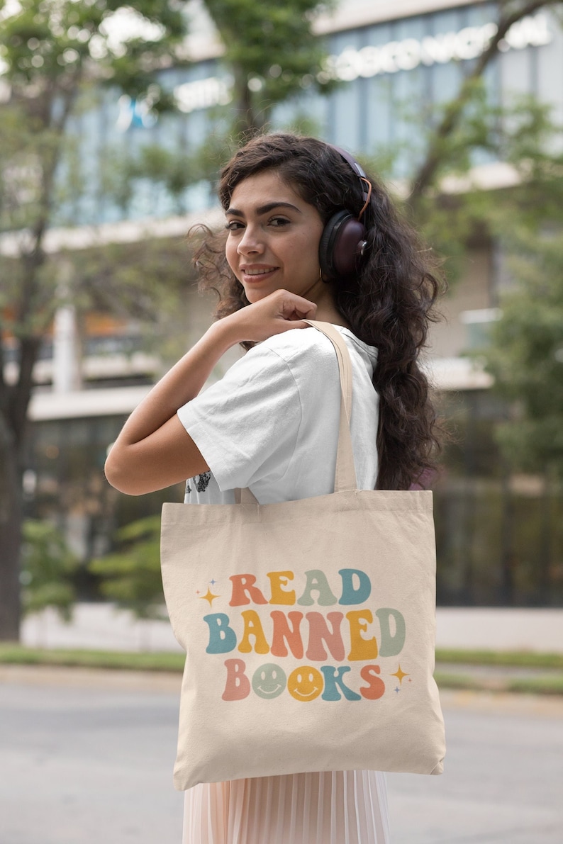 Peut inclure: Une personne portant un sac fourre-tout beige avec le texte "READ BANNED BOOKS" en couleurs arc-en-ciel avec des smileys.