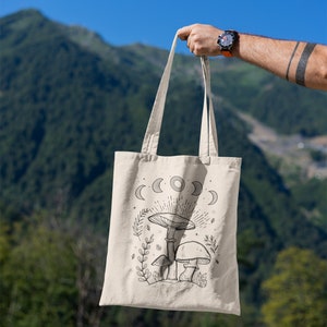 K&ouml;nnte beinhalten: Eine beige Tragetasche mit einer schwarz-wei&szlig;en Illustration von Pilzen und einem Halbmond-Design. Die Tasche wird vor dem Hintergrund einer Bergkette gehalten.