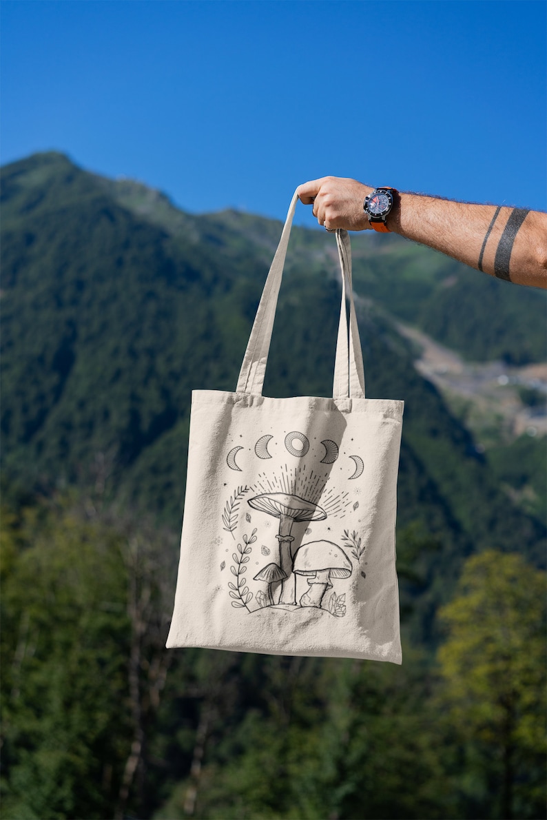 K&ouml;nnte beinhalten: Eine beige Tragetasche mit einer schwarz-wei&szlig;en Illustration von Pilzen und einem Halbmond-Design. Die Tasche wird vor dem Hintergrund einer Bergkette gehalten.