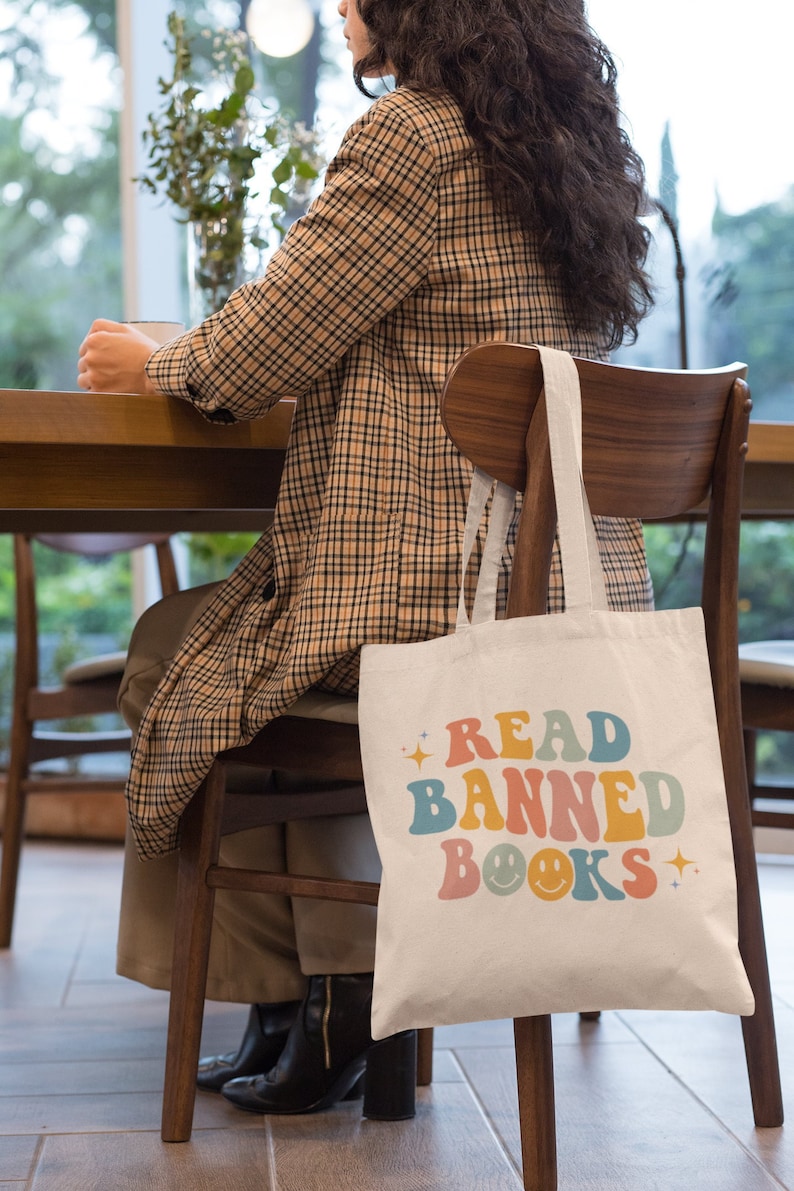 Peut inclure: Un sac en toile blanc avec le texte "READ BANNED BOOKS" en lettres r&eacute;tro color&eacute;es et des smileys. Le sac est accroch&eacute; au dossier d'une chaise en bois.
