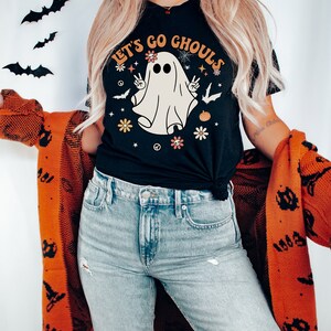 May include: A black t-shirt with a white ghost graphic and the text "Let's Go Ghouls". The ghost has flowers and peace signs around it. The person wearing the shirt is also wearing blue jeans and a red cardigan with black bats.