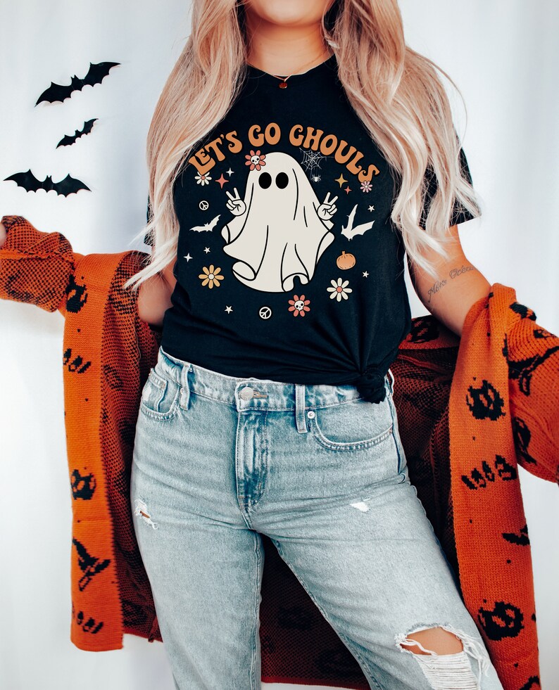 May include: A black t-shirt with a white ghost graphic and the text "Let's Go Ghouls". The ghost has flowers and peace signs around it. The person wearing the shirt is also wearing blue jeans and a red cardigan with black bats.
