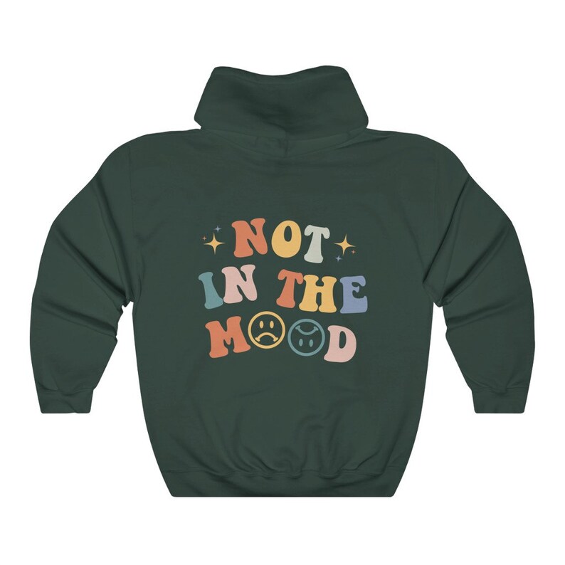Pu&ograve; includere: Una felpa con cappuccio verde scuro con il testo "NOT IN THE MOOD" in colori arcobaleno con emoji di facce tristi.