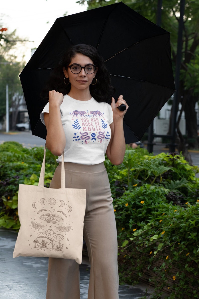 May include: A white tote bag with a brown design of a mushroom, moths, and crescent moons. The bag is being held by a person wearing a white t-shirt with the text "You are made of magic" and a pink and purple tiger design.