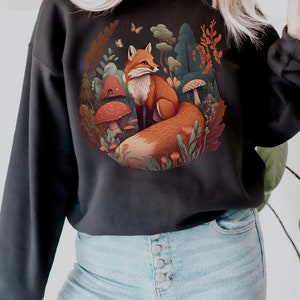May include: A black sweatshirt featuring a graphic print of a fox sitting in a woodland scene. The fox is orange and white, and the woodland is filled with green leaves, mushrooms, and flowers.