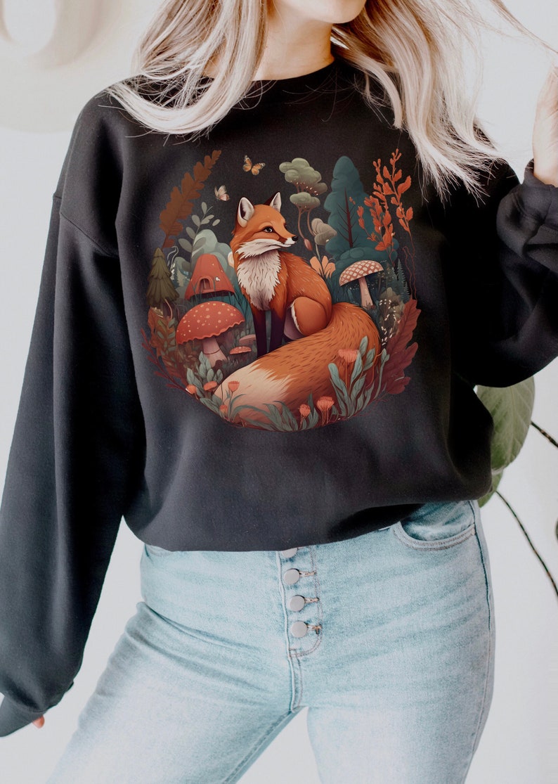 May include: A black sweatshirt featuring a graphic print of a fox sitting in a woodland scene. The fox is orange and white, and the woodland is filled with green leaves, mushrooms, and flowers.