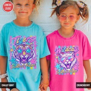 May include: A teal t-shirt with a graphic of a tiger wearing star-shaped sunglasses and the text "Tigers School Name" and a pink t-shirt with a graphic of a cat wearing heart-shaped sunglasses and the text "Wildcats School Name".