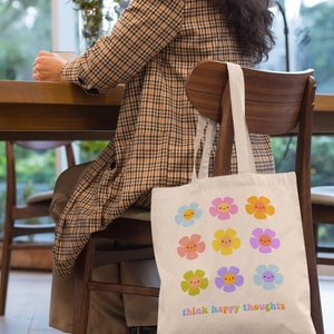 Pu&ograve; includere: Una borsa tote in tela bianca con un design floreale colorato e il testo "think happy thoughts".