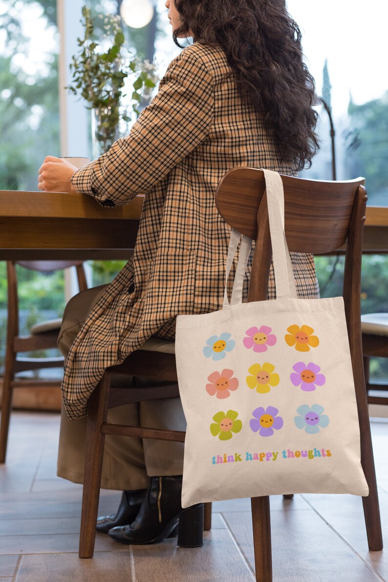 Pu&ograve; includere: Una borsa tote in tela bianca con un design floreale colorato e il testo "think happy thoughts".