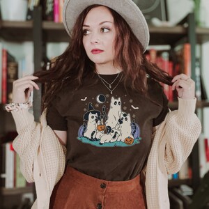 May include: A brown t-shirt with a graphic of four cats dressed up for Halloween. The cats are wearing witch hats, ghost costumes, and holding jack-o'-lanterns. The graphic is set against a dark blue background with stars and bats.