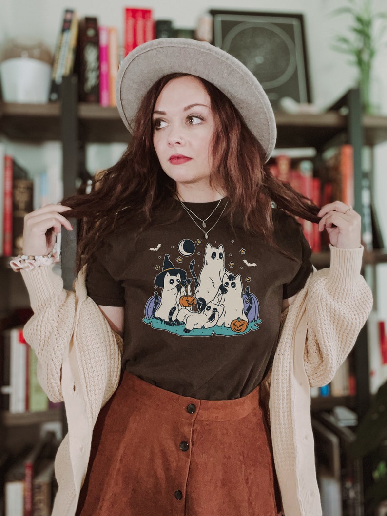 May include: A brown t-shirt with a graphic of four cats dressed up for Halloween. The cats are wearing witch hats, ghost costumes, and holding jack-o'-lanterns. The graphic is set against a dark blue background with stars and bats.