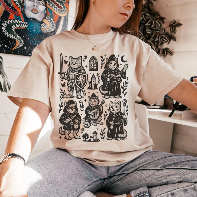 May include: A beige t-shirt with a black and white illustration of five cats dressed as fantasy characters. The cats are a knight, a wizard, a bard, a rogue, and a cleric. The illustration is surrounded by stars, leaves, and a castle.