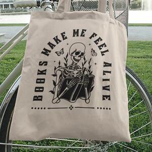 Pu&ograve; includere: Una borsa di tela beige con testo nero e un'immagine di uno scheletro che legge un libro. Il testo dice "I libri mi fanno sentire vivo".
