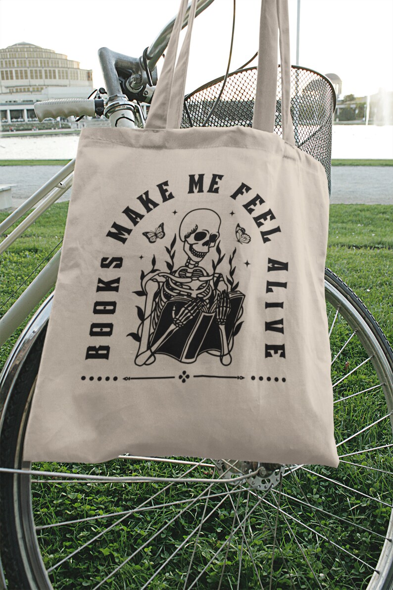Pu&ograve; includere: Una borsa di tela beige con testo nero e un'immagine di uno scheletro che legge un libro. Il testo dice "I libri mi fanno sentire vivo".