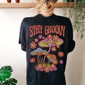May include: Black t-shirt with a graphic of colorful mushrooms and flowers. The text "STAY GROOVY" is printed in red on the shirt.