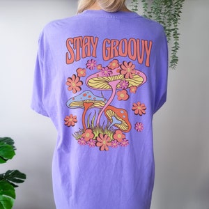 May include: Purple t-shirt with a graphic of mushrooms and flowers. The text "Stay Groovy" is printed on the shirt.