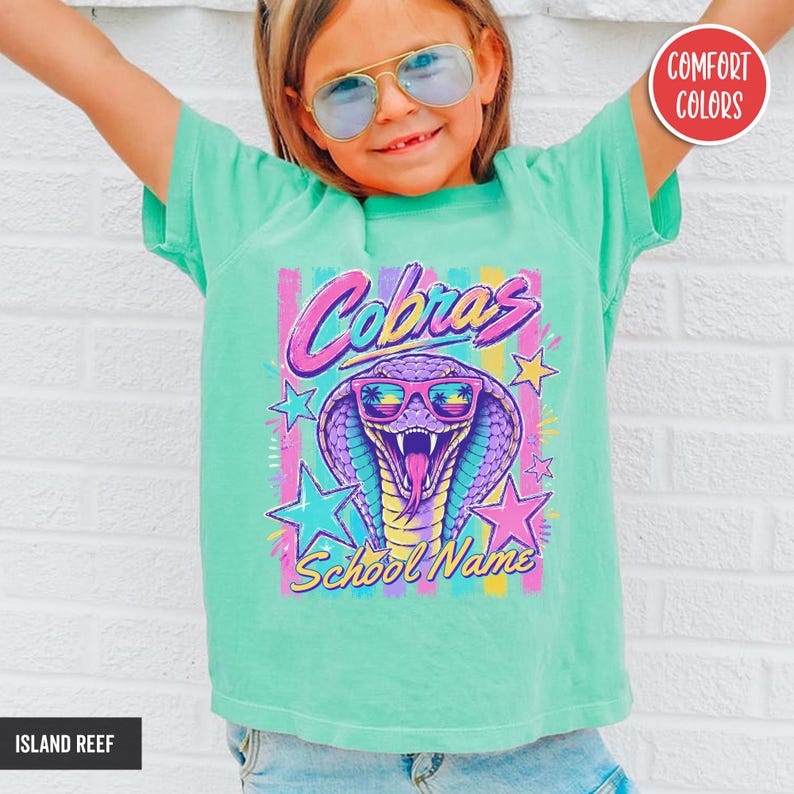 May include: A mint green short-sleeve t-shirt featuring a graphic of a purple cobra wearing pink sunglasses. The text reads Cobras School Name in a stylized font. The design includes colorful vertical stripes and stars in pink, yellow, and blue.