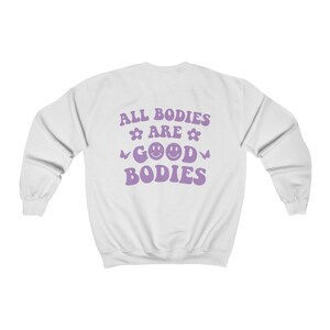 May include: White sweatshirt with the text "ALL BODIES ARE GOOD BODIES" in purple. The text includes flower and butterfly graphics, and two smiley faces. The sweatshirt has long sleeves and a crew neck.